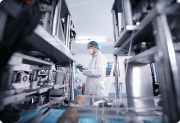 A scientist or technician in protective clothing, including a lab coat, mask, and hairnet, operating machinery in a sterile laboratory or pharmaceutical manufacturing facility.
