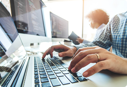 Close-up of a person typing on a laptop keyboard with multiple monitors displaying code, while another person works in the background in a modern office setting.