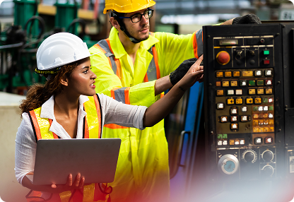 A female engineer holding a laptop collaborates with a male technician in safety gear as they operate a complex industrial control panel in a manufacturing facility.