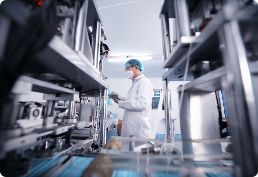 A scientist or technician in protective clothing, including a lab coat, mask, and hairnet, operating machinery in a sterile laboratory or pharmaceutical manufacturing facility.